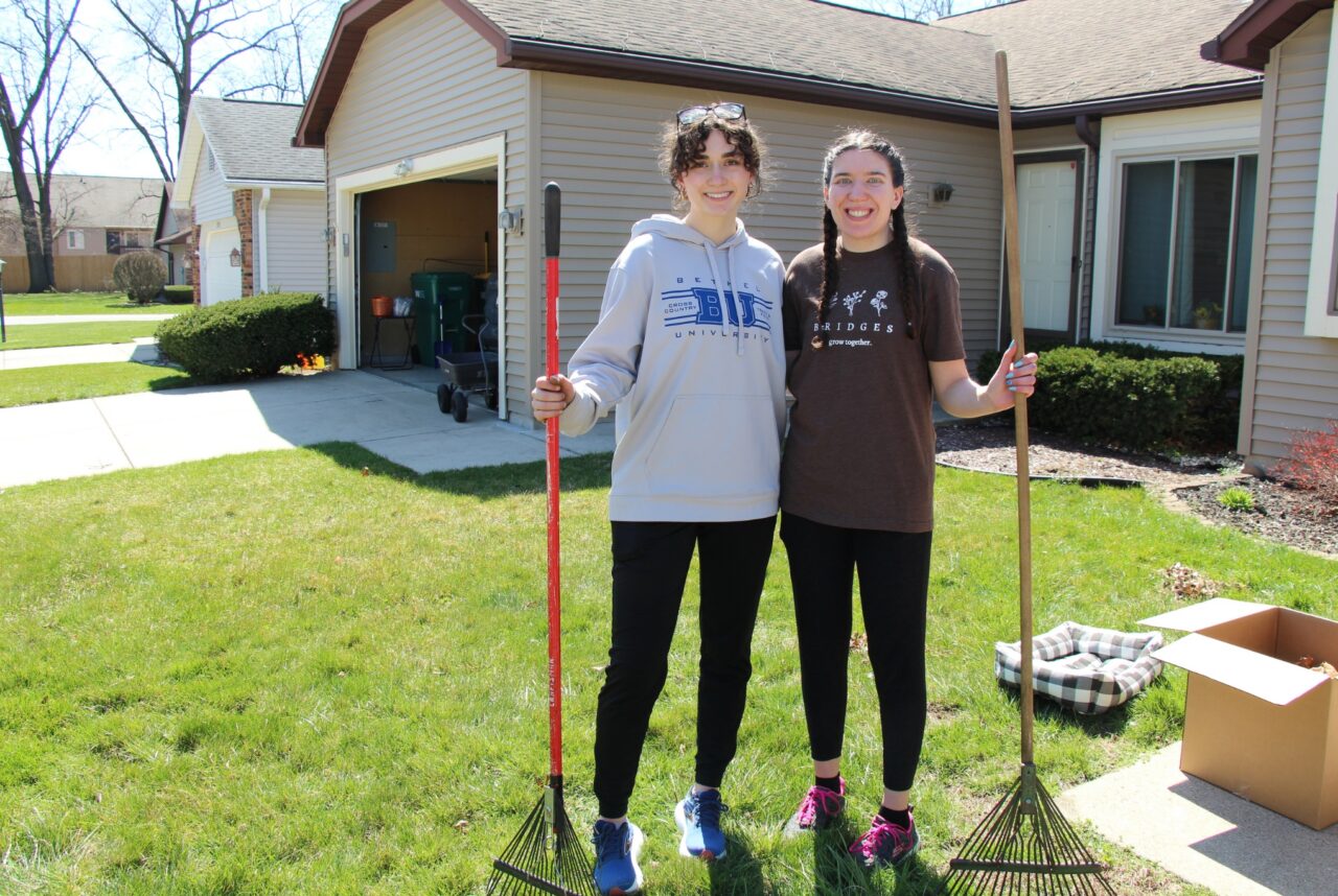 Two young women hold rakes to do yard work in front of house.