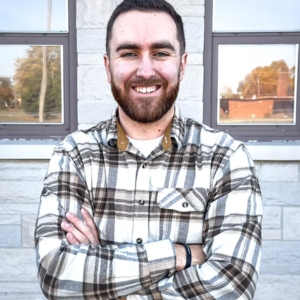 Ryan Lutz ’19, Bethel University’s 2026 Young Alumnus of the Year, smiling with his arms crossed in front of a light gray building.